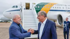 Lula e Alckmin sorrindo e apertando as mãos em frente a avião brasileiro