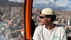 Gabriel, um homem pardo de boné para trás olha a cidade enquanto anda de teleférico