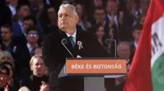 Viktor Orbán - siwowłosy mężczyzna w ciemnym garniturze - stoi na mównicy. Za nim widzimy dużą grupę osób na trybunach, a przed nim - węgierską flagę. 