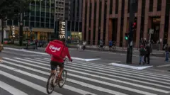 Um entregador anda de bicicleta enquanto faz uma entrega na Avenida Paulista, em São Paulo. Ele atravessa a faixa de pedestre e tem uma mochila vermelha com a marca iFood