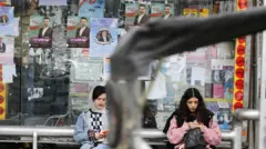 زنی بدون حجاب در کنار زنی با روسری عقب رفته در ایستگاه اتوبوسی نشسته‌اند و مردی در کنار آنها ایستاده است و تبلیغات انتخاباتی پشت آنها دیده می‌شود