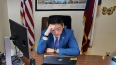 Gene Wu sentado en su escritorio de oficina, mirando un dispositivo frente a él.
