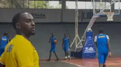 Aristide Mugabe (ibumoso) yambaye umupira w'umuhondo areba ku ruhande, ari mu kibuga kirimo n'abandi bakinnyi benshi ba basketball bambaye amasengeri n'amakabutura by'ubururu. 