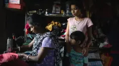 Mulher costurando um tecido vermelho em uma máquina de costura, dentro de um quarto pequeno e escuro, com estantes repletas de frascos e roupas ao fundo. Um menino e uma menina ficam de pé, perto dela.