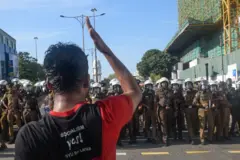 Jovem usando uma camisa preta com vermelha escrita: Sim, socialismo. Na frente dele há uma fila de policiais