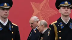Лукашенко і Путін у темних костюмах стоять поруч на тлі великого червоного банера з зіркою. По обидва боки від них — двоє вартових у формі з кашкетами, які стоять нерухомо.