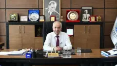 Keşan Belediye Başkanı Mehmet Özcan masasında oturmuş, üzerinde beyaz bir gömlek, arkasında da ödüller, plaketler ve Atatürk resmi var