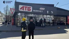 Будівля супермаркету «АТБ», перед входом до якого натягнута червоно-біла стрічка, що обмежує територію. Біля стрічки стоять кілька людей, серед них — рятувальник у захисному спорядженні. На зображенні також є логотип «Суспільне Чернігів».
