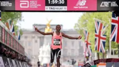 O corredor queniano Sabastian Sawe cruza a linha de chegada da Maratona de Londres de 2026. A imagem mostra o Palácio de Buckingham ao fundo. Acima do corredor, que veste uma blusa rosa e calças justas pretas, encontra-se um cronômetro exibindo o tempo histórico de 1:59:30.