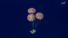 Imagem aérea mostra três paraquedas com listras brancas e vermelhas inflados amortecendo a aterrissagem da cápsula