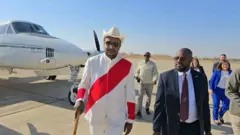 ቦርሳ ኣብ ኢዱ ዝሓዘ ኣምባሳደር ናብ ሑመራ ኣብ ዝኣተውሉ ሓደ ባህላዊ ክዳን ዝለበሰ ብዓልመዚ ኣምሓራ እናተቐበሎ