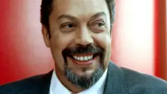 Timy Curry in grey suit in front of red wall