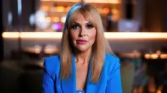 Uma mulher vestindo um blazer azul brilhante sobre uma blusa da mesma cor, sentada em um ambiente interno com iluminação suave ao fundo, apresentando luzes desfocadas e prateleiras.