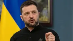 Volodymyr Zelensky. Uyu Perezida wa Ukraine afite ubwanwa, yambaye umupira w'amaboko maremare w'umukara ndetse arimo gukora ikimenyetso n'ikiganza cye cy'ibumoso, mu gihe arimo kuvugira mu nzu, inyuma ye hari ibendera rya Ukraine
