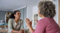 Duas mulheres sentadas a uma mesa. Uma delas levanta as mãos para deter a conversa da outra