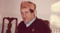 Fotografia de Ronnie Lockwood, que usa um chapéu de papel vermelho típico do Natal. Ele veste uma camisa azul com um suéter azul-marinho com decote em V por cima. Ele tem olhos azuis brilhantes e cabelos castanhos e sorri para a câmera.