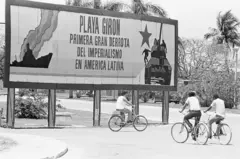 Tres personas en bicicleta pasan junto a un cartel de propaganda que exalta la "derrota del imperialismo" en Playa Girón, Cuba.