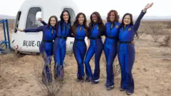 Seis mulheres estão de pé vestindo macacões azuis