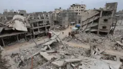 Palestinos caminham junto aos edifícios destruídos em Khan Yunis, no sul da Faixa de Gaza, 22 de janeiro de 2026