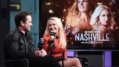 Charles Esten e Hayden Panettiere em um evento para a imprensa com uma placa escrito "Nashville" ao fundo.