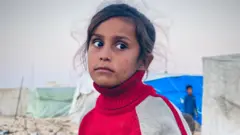 طفلة فلسطينية ترتدي قميصاً أحمر وخلفها خيام، تنتظر دورها للحصول على مياه للشُرب في رفح جنوبي قطاع غزة