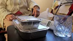 Two foil food containers stacked, and empty glass and a landline wireless phone sit on a black leather-like surface. Behind it is an elderly person wearing a beige cable knit cardigan with buttons. Face is not visible and clothing does not make it possible to distinguish if they are male or female.