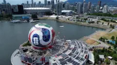 Şehir merkezinin uzaktan ve havadan görüntüsünün içinde maçın oynanacağı stadyum var. En önde de yakın plandaki bir yapının üzerinde dev bir futbol topu var.