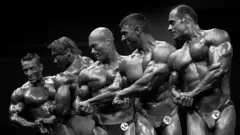 A imagem em preto e branco mostra cinco fisiculturistas alinhados em um palco durante uma competição de fisiculturismo. Vestindo apenas sungas de competição com números presos à frente, eles executam uma pose lateral para destacar braços, peitoral, ombros e abdômen, enquanto os músculos aparecem bastante definidos e iluminados pelos refletores do evento.