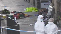 Dos personas, vestidas con trajes de seguridad blancos, se encuentran en un andén. Están detrás de la cinta policial azul y blanca que acordonó una zona llena de basura y equipo de ambulancia.