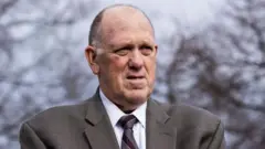 Tom Homan stands outside in front of leafless trees wearing a grey suit.