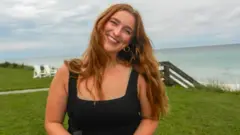 Lizzy'nin uzun, kızıl saçları var ve sahile yakın çimenli bir uçurumun üzerinde dururken siyah bir üst giyiyor. Gökyüzü bulutlu ve arkasında sahile inen ahşap basamaklar ve beyaz şezlonglar görünüyor.