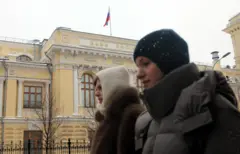 Dwie kobiety w strojach zimowych spacerują chodnikiem przed budynkiem Centralnego Banku Federacji Rosyjskiej. Nad kremowożółtym budynkiem z białymi zdobieniami powiewa rosyjska flaga. Zdjęcie: Maxim Shipenkov / EPA / Shutterstock.
