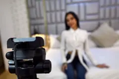 Una muchacha posa ante una cámara web vistiendo una camisa blanca y unos blue jeans. Ella está desenfocada, y está sentada sobre una cama.