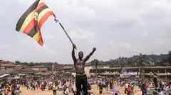 A young shirtless man stands on top of a small mound of dirt, waving the Ugandan flag. Behind him is a crowd of people at a rally led by opposition figure, Robert Kyagulanyi Ssentamu.