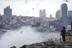 İstanbul Üsküdar'da lodos etkisiyle deniz dalgası kıyıya vurmuş ve martılar havada uçuşuyor, bir kadın kıyıda duruyor, arka planda apartmanlar görünüyor