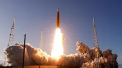 大きな炎を噴き出したロケットが先端に宇宙船をつけて上空へと昇っている。地上では大きな煙が発生して広がっている。周囲には鉄塔がいくつか立っている