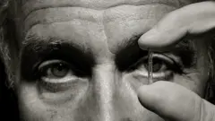 Julio Palmaz en fotografía blanco y negro, primer plano, sólo ojos y sus dedos sosteniendo un stent.