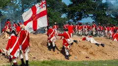 Homens com uniformes vermelhos e brancos sobem uma duna durante uma encenação de batalha