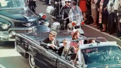 Kennedy üstü açık bir arabada eşiyle birlikte. 