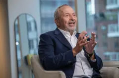 Larry Summers está sentado em uma poltrona cinza, em um apartamento. Atrás dele há um espelho. Ele usa um terno azul e uma camisa branca e gesticula com as mãos enquanto fala.