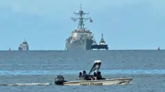O destróier USS Gravely pode ser visto ao fundo da imagem, na costa de Trinidad e Tobago. Mais à frente, há uma lancha do serviço da Guarda Costeira do país.