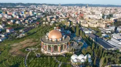 Foto aérea mostra cidade e santuário rodeado por jardins 