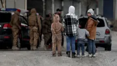 Dört çocuk, arkaları dönük, yalnızca biri kameraya bakıyor. Arka planda iki otomobil ve askeri giyimli kişiler var.