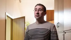 Олег Ролдугин в очках и свитере в полоску на фоне шкафов и открытой двер.