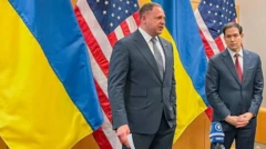 أندري يرماك (يسار)، يرتدي بدلة رسمية رمادية اللون وقميصاً أبيض وربطة عنق رمادية مخططة بالأسود، بجانبه يقف وزير الخارجية الأمريكي ماركو روبيو مرتدياً بدلة رسمية رمادية اللون أيضاً وقميصاً أبيض وربطة عنق تبدو حمراء اللون، يبدو أنهما يتحدثان وخلفهما أربعة أعلام، علمان للولايات المتحدة، وعلمان لأوكرانيا