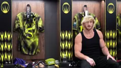 Logan Paul, homem musculoso de cabelo loiro platinado, veste uma regata preta enquanto está sentado em um vestiário
