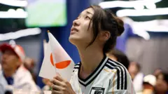 Japon bir kadın taraftar, elinde Japonya bayrağı ve üzerinde japonya üniformasıyla endişeli bir şekilde bir ekrandan maçı izliyor. 