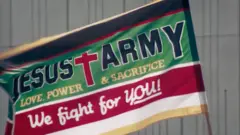 Bandeira do Exército de Jesus, em verde e vermelho, com uma grande cruz vermelha e os dizeres em inglês: 'Amor, Poder e Sacrifício: nós lutamos por VOCÊ!'