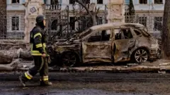 Umuntu uzimya umuriro ari iruhande rw'imodoka yangiritse nyuma y'igitero mu mujyi wa Sumy muri Ukraine 