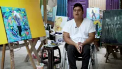 Homem em cadeira de rodas; ao lado dele, há várias pinturas em cores vivas.